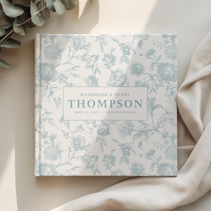 May include: A square, hardcover book with a floral pattern in light blue and white. The cover features the names "Katherine & Henry Thompson" and the date "May 22, 2027 | San Francisco" in a rectangular frame.