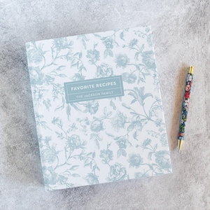 May include: A recipe book with a floral design in shades of blue and white, with the text "Favorite Recipes" and "The Jackson Family". A matching pen with a floral pattern is next to the book. The book is a square shape.
