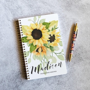 May include: A spiral-bound notebook with a white cover featuring a watercolor sunflower illustration, the name Madison, and the text PASSWORD KEEPER, accompanied by a retractable pen with a floral pattern in shades of pink, white, and black.