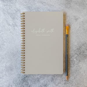 May include: A light gray spiral-bound notebook with gold wire binding. The cover features the white text "elizabeth smith" in a script font above the printed text "LOGINS + PASSWORDS". A black pencil with a gold ferrule and pink eraser is placed beside it.