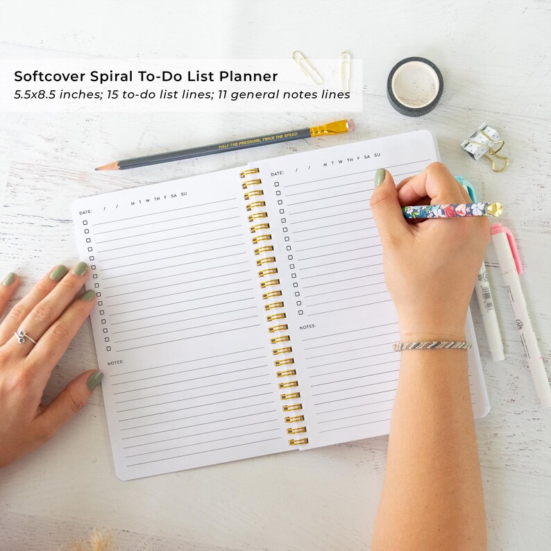 A person's hands are seen writing in a spiral-bound notebook with a 5.5 x 5.5 inch page, which appears to be a softcover spiral to-do list planner. The notebook is placed on a white surface, surrounded by various office supplies such as pens, pencils, and a stapler.
