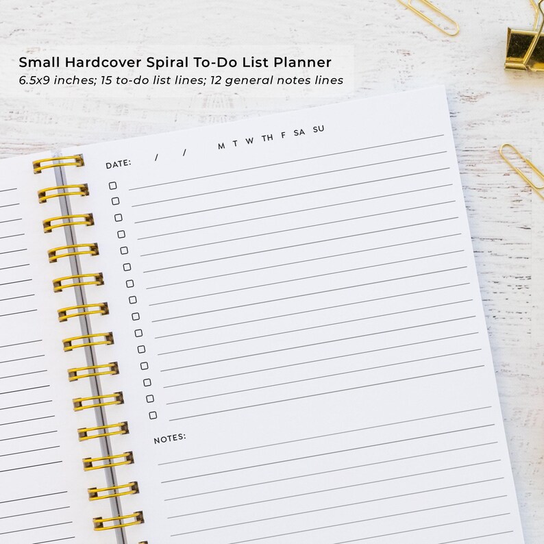 a spiral-bound notebook with a blank page, accompanied by a stapler and a pair of gold binder clips on a wooden surface.
