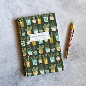 May include: A spiral-bound notebook with a dark green cover featuring a repeating pattern of potted plants in various colors. A gold pen with a floral design rests beside the notebook. The notebook has the text "GRACE O'DONNELL tasks and notes".