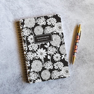 May include: A spiral-bound password notebook with a black cover featuring a white line-art floral pattern. A rectangular label in the center displays the text PASSWORDS and Rachel Norlin. A gold-tone pen with a colorful floral pattern is placed beside it.