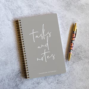 May include: A grey spiral-bound notebook with the words "tasks and notes" in white script. A floral patterned pen with a gold barrel rests beside the notebook. The notebook is labelled "Chandler Peterson".