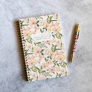 May include: A floral notebook and pen set. The notebook features a cream background with pink flowers and green leaves. The pen has a gold barrel and a floral design. The notebook reads "Olivia Gonzalez TASKS + NOTES".