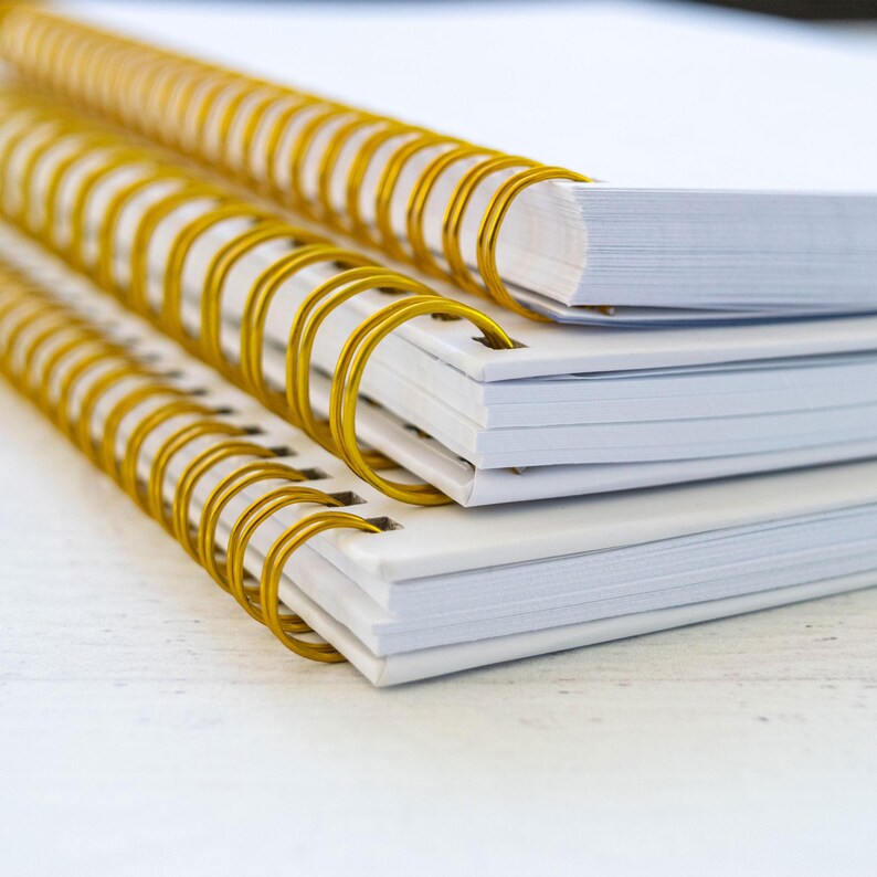 A stack of white spiral-bound notebooks with gold spiral bindings.