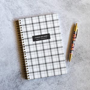May include: A spiral-bound notebook with a white and gray plaid pattern featuring a black rectangular label that reads CHRIS NELSON logins + passwords, accompanied by a retractable pen with a floral pattern and gold-toned metal accents.