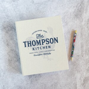 May include: A white notebook with a blue and white logo that says "The Thompson Kitchen" with the text "Established 2020" and "Fresh Eats - Lively Conversation" and "Lexington, Kentucky".