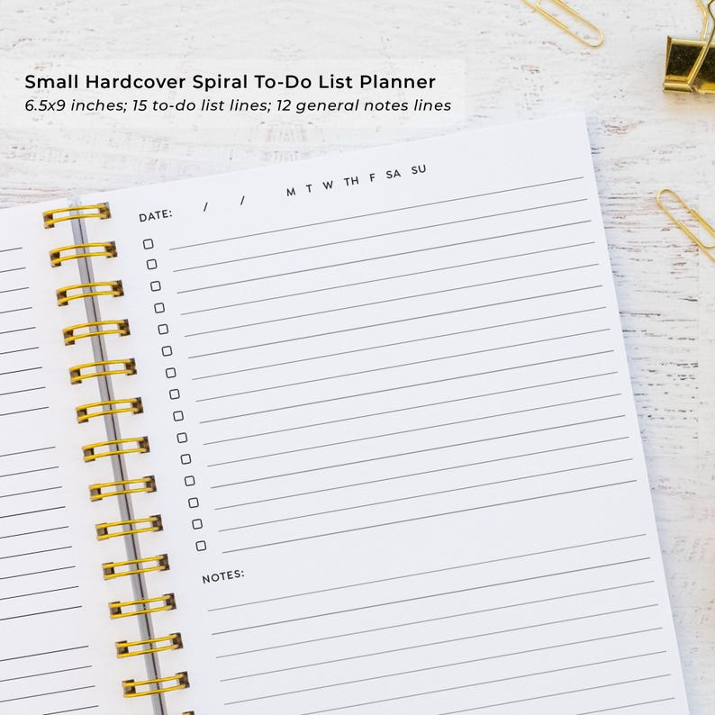 a spiral-bound notebook with a blank page, accompanied by a stapler and a pair of gold binder clips on a wooden surface.