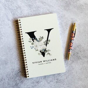 May include: A cream-colored spiral notebook with a large black letter "V" adorned with white and blue floral accents. The notebook is next to a pen with a floral pattern and gold accents. The text "VIVIAN WILLIAMS TASKS + NOTES" is printed on the notebook.