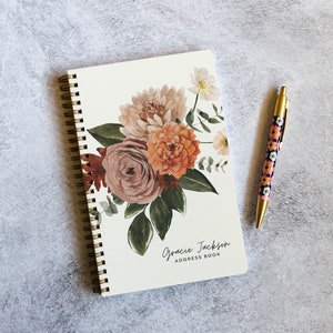 May include: A white spiral bound address book with a floral design featuring pink, orange, and green watercolor flowers. The text "Gracie Jackson ADDRESS BOOK" is printed on the cover.