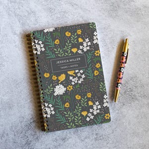 May include: A notebook with a floral pattern in yellow, white, and green on a dark grey background. The cover reads "JESSICA MILLER TASKS + NOTES". A pen with a floral design and gold accents lies next to the notebook. The notebook is spiral-bound.