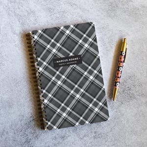 May include: A spiral-bound notebook featuring a gray, black, and white diagonal plaid pattern with a central black rectangular label reading MARCUS ADAMS logins + passwords, accompanied by a gold-toned pen with a floral pattern of pink and white flowers.
