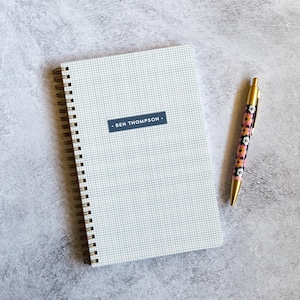 May include: A spiral-bound notebook with a white cover featuring a black grid pattern and a dark blue rectangular label printed with the name BEN THOMPSON. Beside the notebook lies a gold-toned pen with a floral pattern of pink and white flowers.