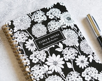 May include: A spiral-bound notebook with a black cover featuring a white floral pattern. The notebook has a gold spiral binding and a rectangular label that reads "TASKS & NOTES" and "Rachel Norlin". A black and silver pen rests on the side.