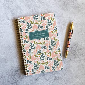 May include: A spiral-bound notebook with a floral and heart pattern in shades of pink, green, and white. A rectangular teal label in the center reads Erin Williams LOGINS + PASSWORDS. A matching retractable pen with a floral print is included.
