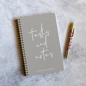 May include: A gray spiral bound notebook with the text "tasks and notes" in white lettering. The notebook has the name "Chandler Peterson" printed on the bottom. A gold pen with a floral pattern is lying on a gray surface next to the notebook.