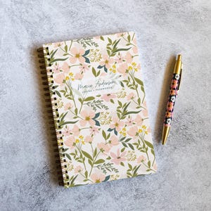 May include: A spiral-bound notebook with a cream background featuring a pattern of pink flowers, green leaves, and yellow sprigs. A white rectangular label in the center reads Macie Anderson LOGINS + PASSWORDS. A matching floral pen sits beside it.