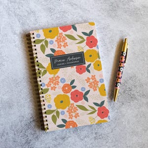 May include: A spiral-bound notebook with a light pink cover featuring a floral pattern of yellow, orange, and red flowers with green leaves. A black rectangular label in the center reads Macie Anderson LOGINS + PASSWORDS. A matching floral pen.