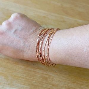 Peut inclure: Plusieurs bracelets fins en cuivre martel&eacute; empil&eacute;s sur un poignet. Les bracelets ont une surface textur&eacute;e et une couleur brun-rouge&acirc;tre chaude. L'arri&egrave;re-plan est une surface en bois de couleur claire.