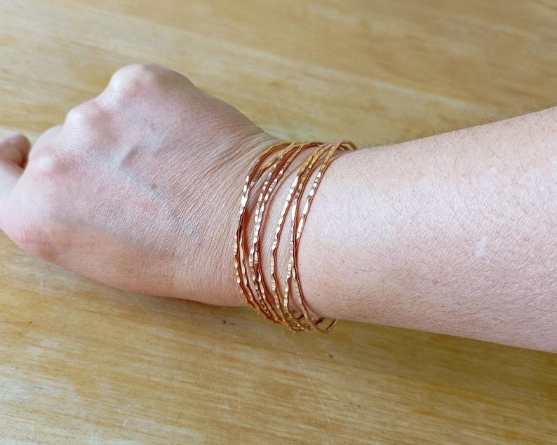 Peut inclure: Plusieurs bracelets fins en cuivre martel&eacute; empil&eacute;s sur un poignet. Les bracelets ont une surface textur&eacute;e et une couleur brun-rouge&acirc;tre chaude. L'arri&egrave;re-plan est une surface en bois de couleur claire.