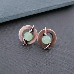 Puede incluir: Un par de pendientes de cobre con una piedra verde en el centro de cada pendiente. Los pendientes están hechos de alambre envuelto alrededor de un círculo de cobre.