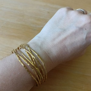 May include: A gold-toned bracelet made of multiple thin, hammered metal bands. The bracelet is worn on a person's wrist.