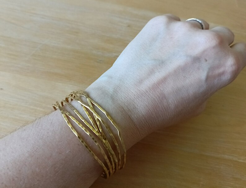 May include: A gold-toned bracelet made of multiple thin, hammered metal bands. The bracelet is worn on a person's wrist.
