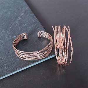 Peut inclure: Deux bracelets cuivr&eacute;s. L'un est un jonc avec plusieurs bandes textur&eacute;es. L'autre est une pile de bracelets fins et textur&eacute;s. Les bijoux sont pr&eacute;sent&eacute;s sur une surface gris fonc&eacute;.