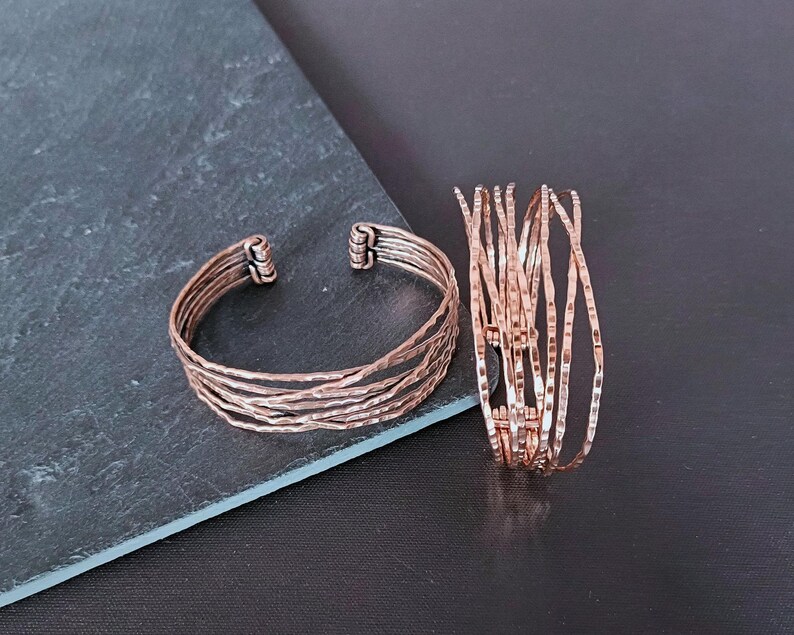 Peut inclure: Deux bracelets cuivr&eacute;s. L'un est un jonc avec plusieurs bandes textur&eacute;es. L'autre est une pile de bracelets fins et textur&eacute;s. Les bijoux sont pr&eacute;sent&eacute;s sur une surface gris fonc&eacute;.