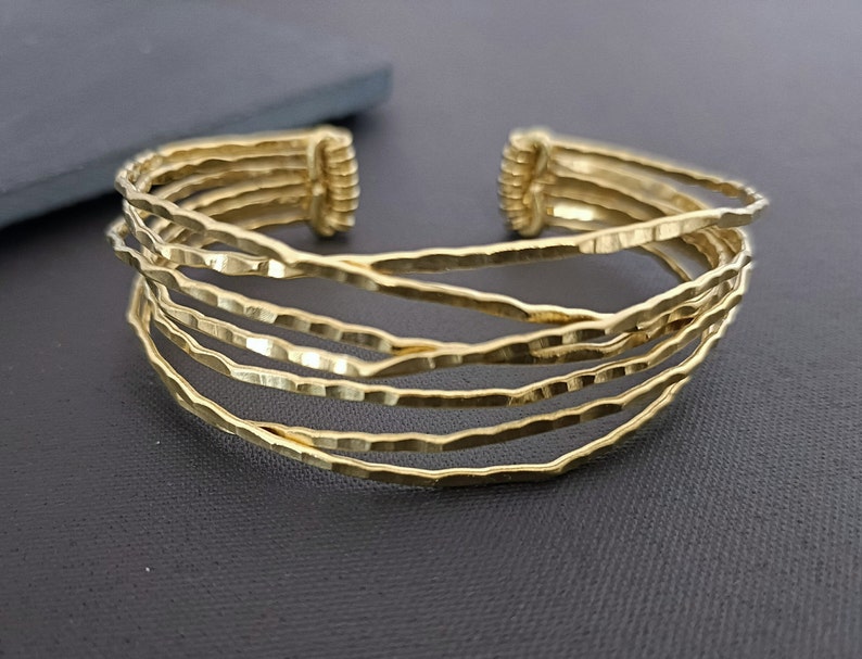 May include: A gold-toned multi-strand cuff bracelet with a hammered texture. The bracelet is made of five thin bands of metal that are woven together.