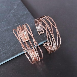 Peut inclure: Deux bracelets en m&eacute;tal couleur cuivre. L'un est de style manchette, l'autre est un mod&egrave;le &agrave; plusieurs brins. Les bracelets ont une texture martel&eacute;e et sont pr&eacute;sent&eacute;s sur une surface gris fonc&eacute;. Le bracelet manchette mesure environ 5 cm de large.
