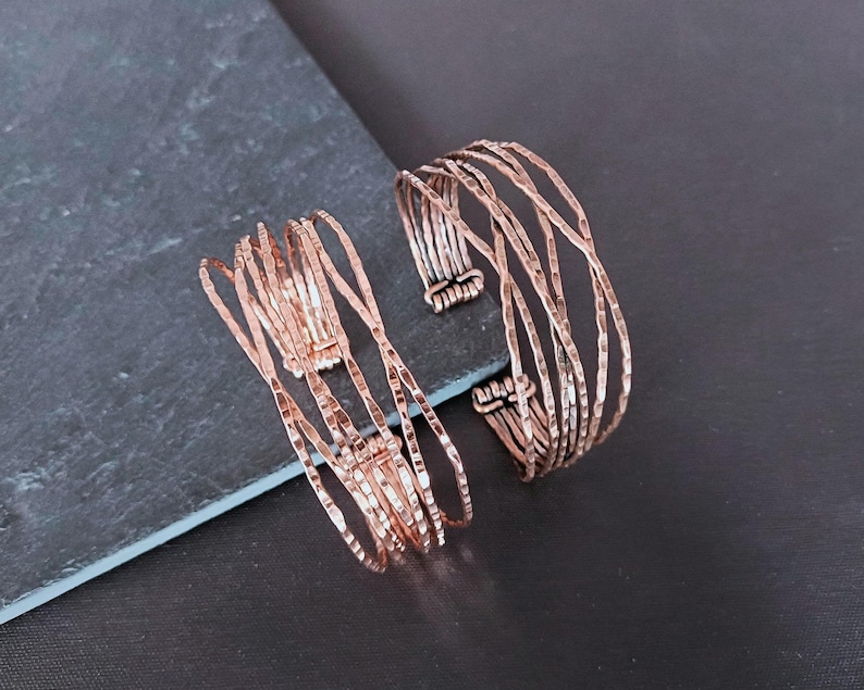 Peut inclure: Deux bracelets en m&eacute;tal couleur cuivre. L'un est de style manchette, l'autre est un mod&egrave;le &agrave; plusieurs brins. Les bracelets ont une texture martel&eacute;e et sont pr&eacute;sent&eacute;s sur une surface gris fonc&eacute;. Le bracelet manchette mesure environ 5 cm de large.