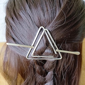 May include: Silver triangle hair clip with a thin bar that goes through the triangle. The clip is in a person's hair.
