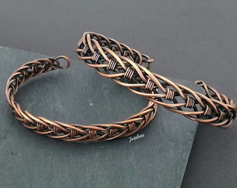 May include: Two copper-colored woven bracelets. One is a closed circle, and the other is an open cuff. The bracelets have a textured, braided design. The bracelets are displayed on a dark gray surface.