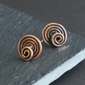 Può includere: Un paio di orecchini a bottone color rame. Ogni orecchino presenta un design a spirale, con una spirale più piccola al centro. Gli orecchini sono appoggiati su una superficie grigio scuro. La parola "Joinhas" è visibile.
