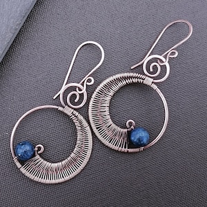 以下が含まれることがあります： 青い宝石のビーズがついた、銀色のワイヤー製のイヤリングのペア。イヤリングは、ワイヤーを自身に巻き付けて円形にしたものです。ワイヤーはねじって螺旋状のデザインになっています。