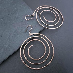 Könnte beinhalten: Ein Paar kupferfarbene Ohrringe mit einem großen, flachen Spiral-Drahtdesign. Jeder Ohrring besteht aus einem einzigen durchgehenden Metallstück, das sich nach innen zu drei konzentrischen Kreisen windet und mit einem einfachen Haken versehen ist.