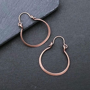 May include: A pair of copper hoop earrings with a rounded, circular design and a simple wire closure mechanism. The earrings feature a warm, metallic reddish-brown finish and a smooth, consistent texture throughout the entire circular frame.