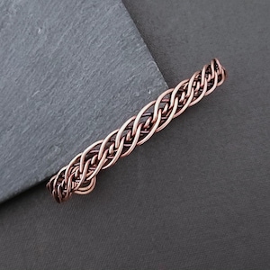 Peut inclure: Un bracelet en métal tressé de couleur cuivre. Le bracelet est composé de brins entrelacés, créant un aspect texturé et tissé. Le bijou est présenté sur une surface gris foncé.