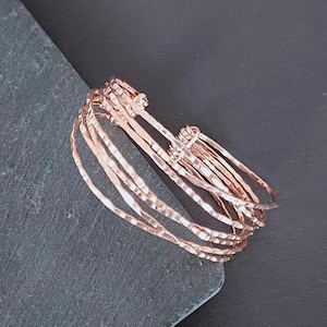 Peut inclure: Une pile de bracelets joncs en or rose avec une texture martel&eacute;e. Les bracelets sont empil&eacute;s, cr&eacute;ant un effet superpos&eacute;. Les bracelets sont pr&eacute;sent&eacute;s sur une surface gris fonc&eacute; sur fond noir.