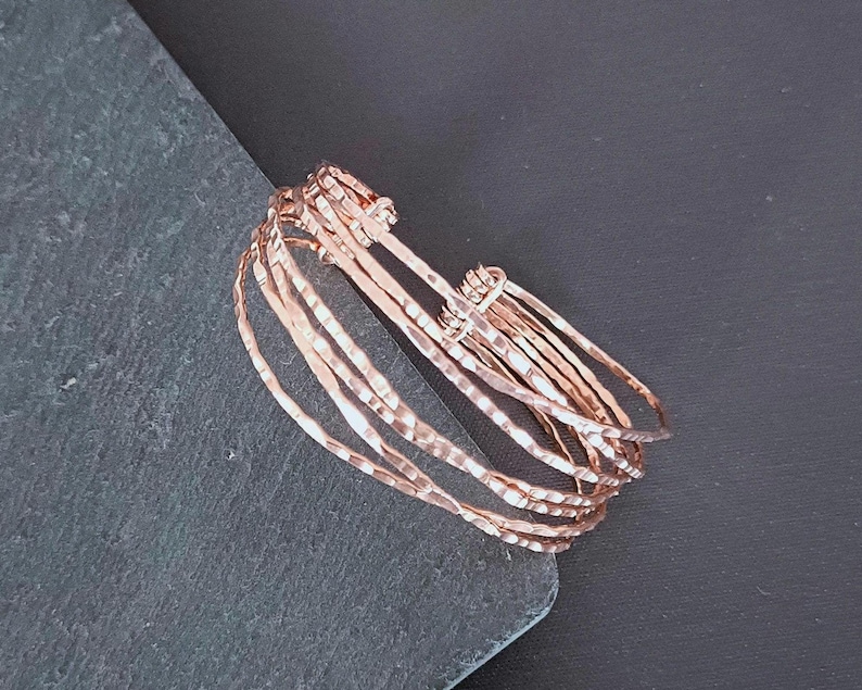 Peut inclure: Une pile de bracelets joncs en or rose avec une texture martel&eacute;e. Les bracelets sont empil&eacute;s, cr&eacute;ant un effet superpos&eacute;. Les bracelets sont pr&eacute;sent&eacute;s sur une surface gris fonc&eacute; sur fond noir.