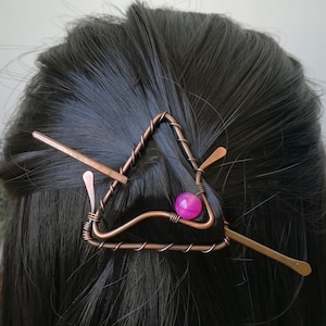 May include: A copper wire hair clip in the shape of a triangle with a pink gemstone in the centre. The clip is being used to hold a woman's hair in a bun.