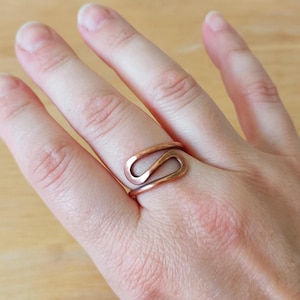 Könnte beinhalten: Ein kupferfarbener Ring mit einem einzigartigen, fließenden Design. Der Ring wird an einem Finger getragen und zeigt seine komplizierte, handgefertigte Form. Das Metall hat einen warmen, rötlich-braunen Farbton, der den künstlerischen Reiz des Rings hervorhebt.