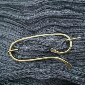 Peut inclure: Épingle à châle dorée avec un design incurvé et une fermeture à épingle droite. L'épingle est présentée sur un tissu gris texturé.