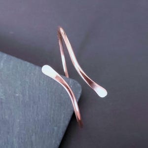 Pode incluir: Um anel aberto em tom de ouro rosa com uma banda de metal fina, lisa e ligeiramente curva que se enrola num design aberto. As extremidades do anel são arredondadas e achatadas, exibindo um acabamento metálico polido em toda a peça.