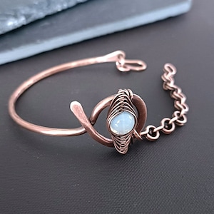 Puede incluir: Una pulsera de cobre con una cadena delicada y una piedra de ópalo blanco engastada en un diseño de alambre envuelto.