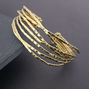 May include: A gold-toned bracelet made of multiple thin, hammered metal bands. The bands are stacked and slightly offset, creating a textured and layered look.