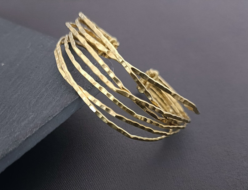 May include: A gold-toned bracelet made of multiple thin, hammered metal bands. The bands are stacked and slightly offset, creating a textured and layered look.
